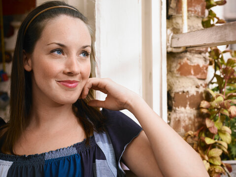 Woman sitting by a window, smiling thoughtfully with her hand on her chin. Germany