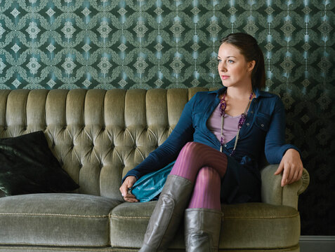 Woman sitting on a vintage sofa against patterned wallpaper, looking thoughtfully to the side. Germany