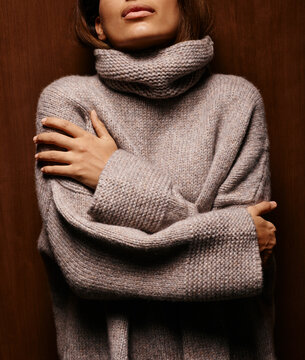 Woman in a cozy knit beige sweater hugging herself against a wooden background. Germany