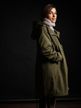 Woman in green coat with scarf poses against a dark background, looking thoughtful. Germany