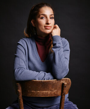 Woman in blue sweater sits confidently on a wooden chair against a dark background. Germany
