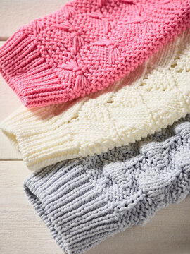 Stacked knitted sweaters in pink, cream, and gray patterns on a light wooden surface. Germany