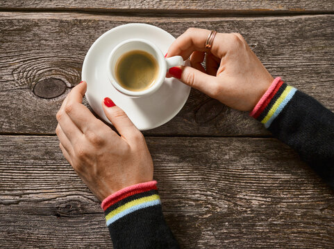 Hands holding an espresso cup on a wooden table with colorful sweater cuffs. Germany