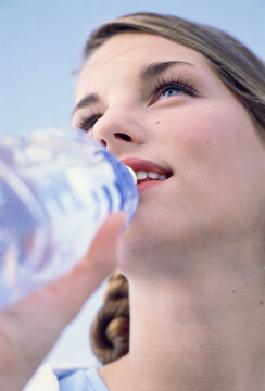 Woman with braided hair drinking from a clear water bottle against a blue sky. Germany