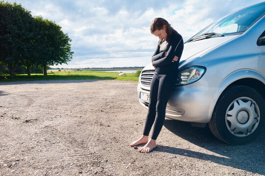 Woman stands barefoot, leaning on a silver van, in a rural setting under a cloudy sky. Baltic Sea, Germany