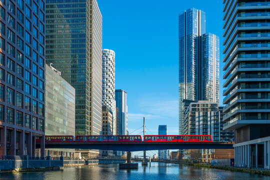 Red train on elevated track amidst tall buildings under a clear blue sky. Canary Wharf, London, UK