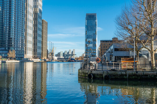 City skyline with skyscrapers reflected in a calm river under a clear blue sky. Canary Wharf, London, UK