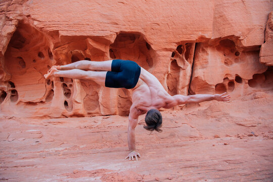 A person balances on one hand on red rock formations with holes under clear daylight. WA, USA