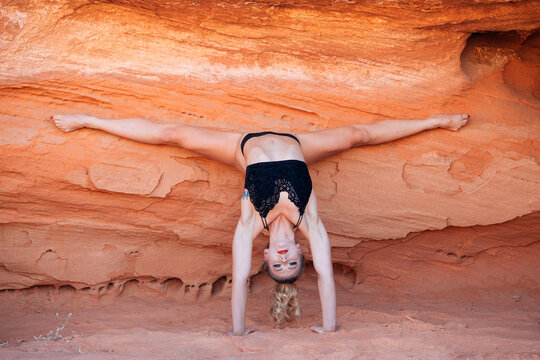 Woman performs a handstand against a red rock wall with legs in a wide split. WA, USA