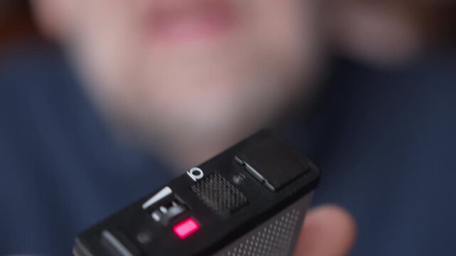 Out-of-focus close-up of a person speaking into the red tally light of a vintage voice recorder. Visual metaphor for testimony and surveillance.