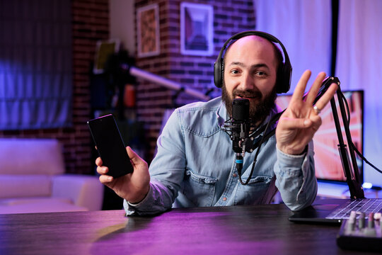 Portrait of tech vlogger presenting three tips for finding smartphones with best value on the market in rgb lights home studio. Content creator filming video, showing counting gesture with fingers