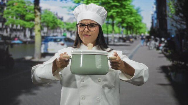 Young woman chef in uniform and toque holding green pot with both hands outstretched on street, wearing glasses and frowning; frustration challenge.