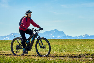 Obraz premium Middle aged woman cycling on an electric bike across green spring meadow in Upper Bavaria with alpine mountain range and Zugspitze silhouette in background under clear blue sky.