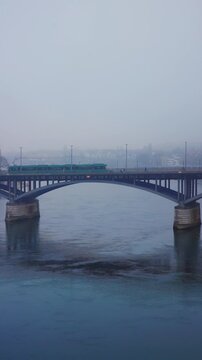 Vertical static shot of a green tram crossing the Wettsteinbruecke bridge over the Rhine river on a misty morning in Basel.