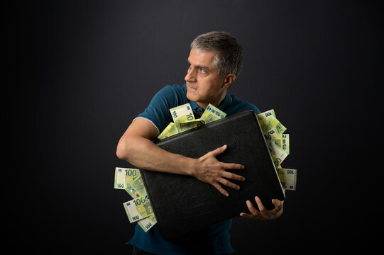 Man holding a suitcase overstuffed with money in a greedy, thievish manner and looking apprehensively over his shoulder. A finance concept with euro banknotes. 