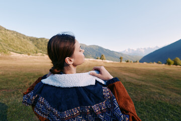 Naklejka premium Woman in patterned jacket standing in meadow outdoors, back portrait gazing at mountain range, autumn light, serene landscape, nature travel and contemplative moment.