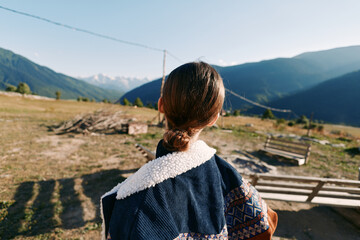 Naklejka premium woman back in jacket with tied hair and shearling collar standing in a meadow by a bench and swing, overlooking mountains and countryside landscape under warm sunlight and open horizon