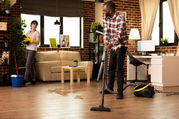 Smiling african american man vacuums hardwood floor while Caucasian wife wipes shelves in modern...