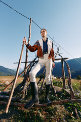 Naklejka premium Woman in rubber boots holds a wooden stick standing by a rustic fence in countryside, wearing patterned jacket and white trousers, posing confidently against mountains and clear sky.