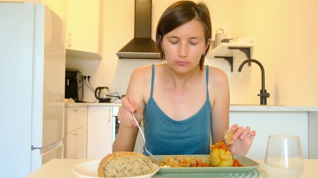 Woman sits at kitchen table eating stuffed paprika, enjoying homemade Balkan cuisine, turning meal into cultural moment shared in quiet domestic space.