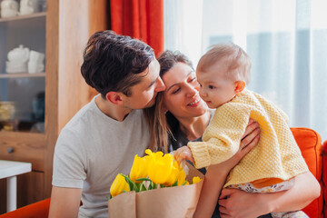 Obraz premium Happy Mother's day. Young man giving his wife bouquet of flowers at home. Woman holding toddler baby girl. Loving parents 