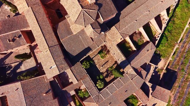 Aerial view shows a building complex with many tiled roofs arranged around a central courtyard with plants in a sunny setting. The layout is geometric and structured.