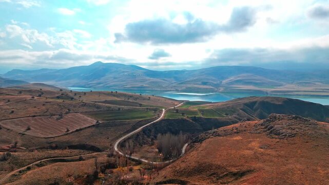 Cinematic aerial view of a winding road, dry hills, distant mountains, and a calm lake under dramatic clouds. A striking rural landscape with natural textures, open space, and travel appeal.