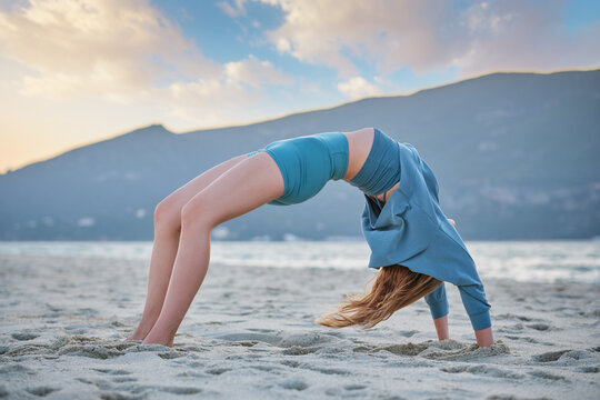 A woman practices Urdhva Dhanurasana on sandy beach at sunset with mountains in the background. She focuses on her yoga pose.