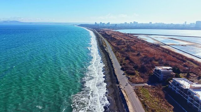 A shoreline stretches along the water with waves rolling in. A road runs parallel to the beach with buildings in the distance. The sky is clear and the water is bright blue.