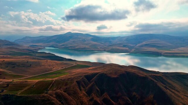 Cinematic aerial footage of a calm lake surrounded by arid hills, farmland, and distant mountains under dramatic clouds. Scenic drone views with reflections, winding roads, and expansive rural terrain