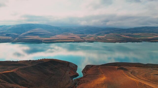 Cinematic aerial footage of a calm lake surrounded by arid hills, farmland, and distant mountains under dramatic clouds. Scenic drone views with reflections, winding roads, and expansive rural terrain