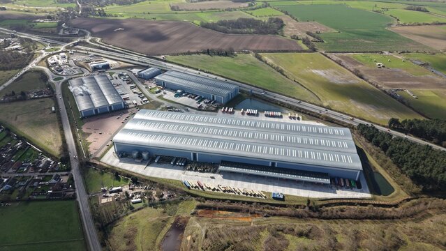 Aerial view of industrial warehouses glinting under the diffused sunlight, contrasting against the surrounding green fields and roadways, Doncaster, Doncaster, United Kingdom.