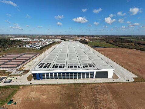 Aerial view of a vast warehouse with a sleek facade and rows of skylights, contrasting with the surrounding brown fields and distant cityscape, Doncaster, United Kingdom.