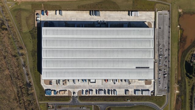 Aerial view of a vast, grey warehouse complex with rows of parked vehicles casting shadows on the tarmac, Doncaster, United Kingdom.