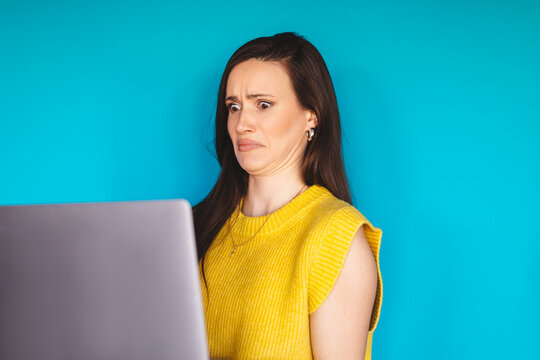 Disgusted young woman looking at laptop screen with grimace, brunette female model feeling aversion or dislike while watching online content on blue background, negative reaction and phobia concept
