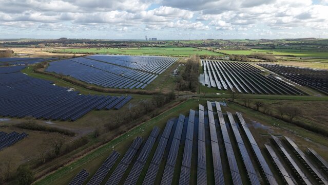 Aerial view of orderly solar panels reflecting the sky's hue, contrasting with the verdant fields and distant structures, Retford, United Kingdom.