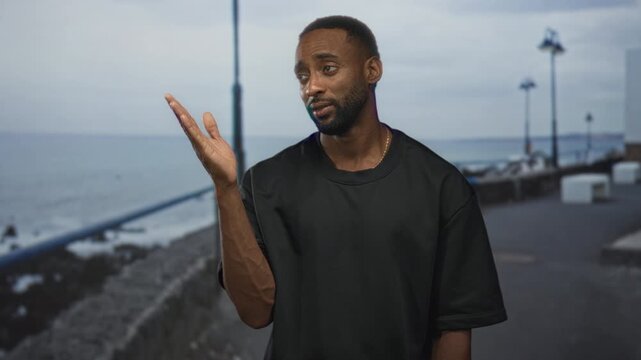 Man pointing finger up with visible forearm on a seaside street promenade near railing and lampposts, wearing black tee and gold chain; thoughtful reflection.