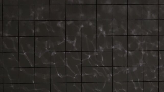 Dark tiled wall illuminated by soft shimmering light reflections forming subtle wave-like patterns across the grid surface, creating a moody abstract texture with ample copy space.