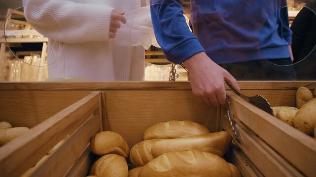 In the bakery section, an anonymous couple uses provided tongs to pick up fresh buns, examining them briefly before adding them to a paper bag held by one of them.