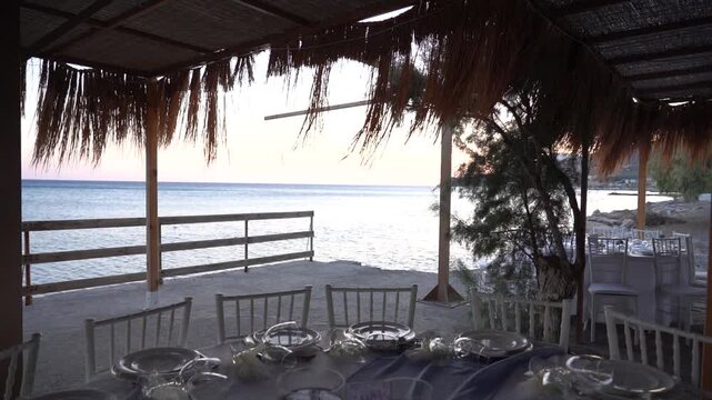 Slow-motion close-up of a straw umbrella gently moving in the breeze above a round table adorned with decorative items on the beach, with sparkling sea in the background.