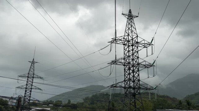 Tall power transmission towers stand against cloudy mountain sky carrying multiple electrical lines across a landscape of hills and distant terrain. Energy infrastructure.