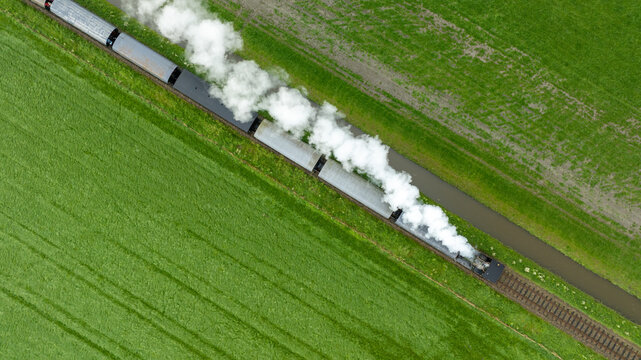 Aerial view of a vintage steam train billowing white smoke as it travels through vibrant green fields, Broerdijk, Benningbroek, Noord-Holland, Netherlands.
