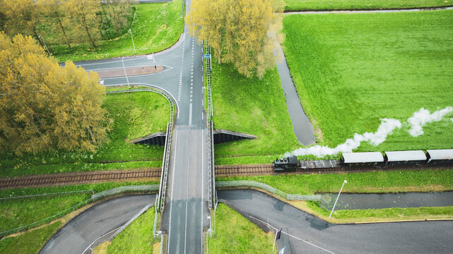 Aerial view of a vintage steam train puffing smoke as it crosses a railway bridge amidst lush green fields and roads, Benningbroek, Noord-Holland, Netherlands.