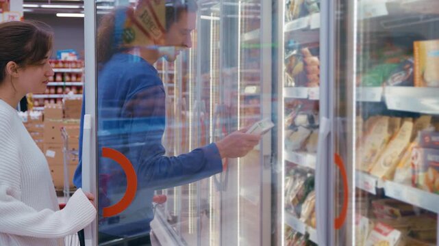 A man and woman with a shopping cart browse the frozen food aisle, opening freezer doors to pull out products for closer inspection. 