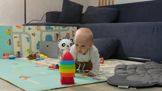 Cute baby boy crawling on a play mat in a living room, reaching for a colorful rainbow rocket toy and trying to chew on it. Toddler learning to crawl, playing, and exploring at home