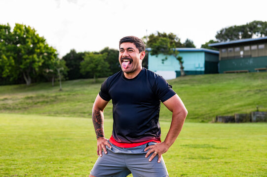 Samoan man doing haka dance standing with arms on his waist sticking his tongue out in grass field