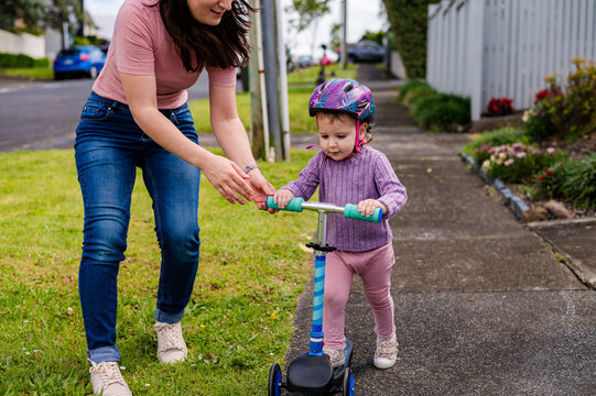 Mum guiding toddler daughter in riding her scooter outdoors