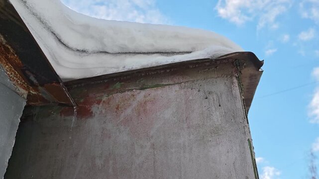 Snow accumulation on metal roof edge melts and drips down the side of a weathered building under a bright blue sky with scattered clouds