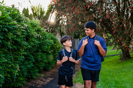 Two boys walk side by side on a path surrounded by green plants while talking to each other