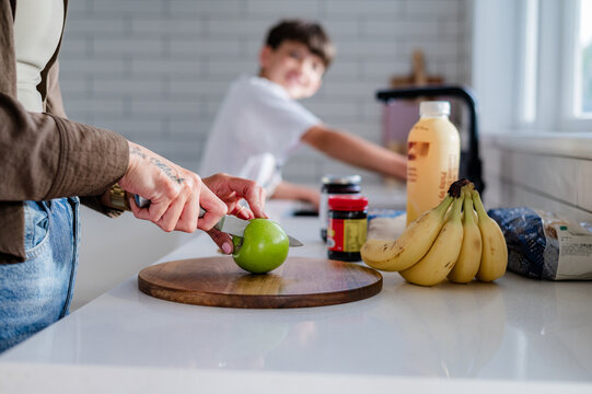 Adult hand cutting an apple on kitchen counter while young boy wash dishes in the background